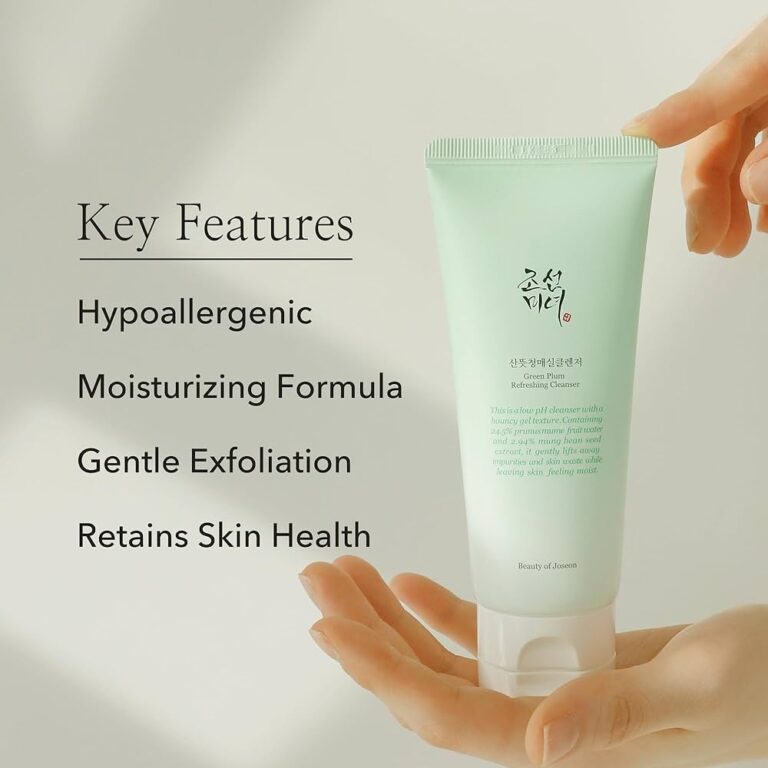 En person holder en grøn tube med Beauty Of Joseon - Green Plum Refreshing Cleanser 100ml med koreansk tekst. Ved siden af fremhæver den beige baggrund den allergivenlige, fugtgivende formel, den blide eksfoliering og hudens sundhedsmæssige fordele.