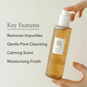 En hånd holder en flaske Beauty Of Joseon - Ginseng Cleansing Oil med koreansk tekst på etiketten. De vigtigste egenskaber er: Fjerner urenheder, renser forsigtigt porerne, har en beroligende duft og efterlader en fugtgivende finish.