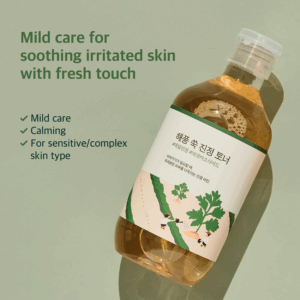En flaske Round Lab - Mugwort Calming Toner 300ml hviler på en grøn overflade med teksten: "Mild pleje, der beroliger irriteret hud med et frisk touch." Højdepunkter: Mild pleje, beroligende virkninger, velegnet til følsom eller kompleks hud.