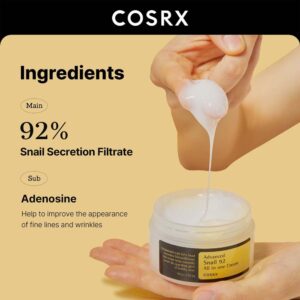 En hånd øser rig hvid creme fra en åben COSRX - Advanced Snail 92 All In One Cream-krukke, der viser sit 92% sneglesekretionsfiltrat og adenosin for anti-aging-fordele.