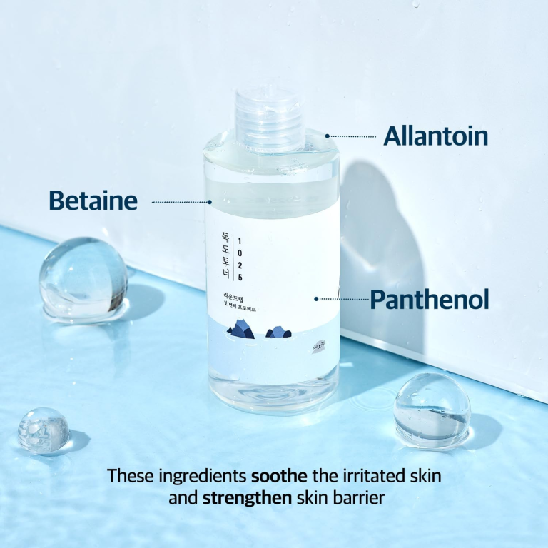 En klar flaske Round Lab - 1025 Dokdo Toner 200ml hviler på en blå overflade med vanddråber. Teksten fremhæver ingredienserne - allantoin, betain og panthenol - og bemærker deres lindrende og styrkende fordele for huden.