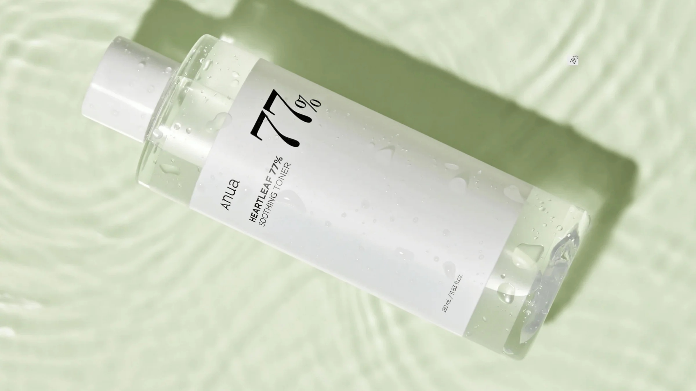 En klar flaske Anua Heartleaf 77% Soothing Toner ligger på en lysegrøn, bølget vandoverflade, der ligner en beroligende header-skabelon. Flasken har en hvid etiket med sort tekst og fine vanddråber.