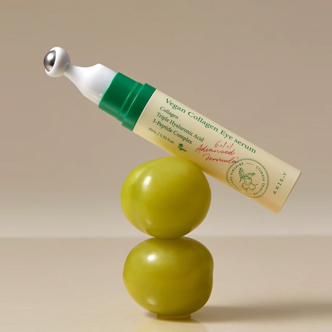 En tube med vegansk kollagenøgeserum med en metalapplikator balancerer på to stablede grønne tomater mod en beige baggrund, hvilket skaber en visuelt slående header-skabelon.