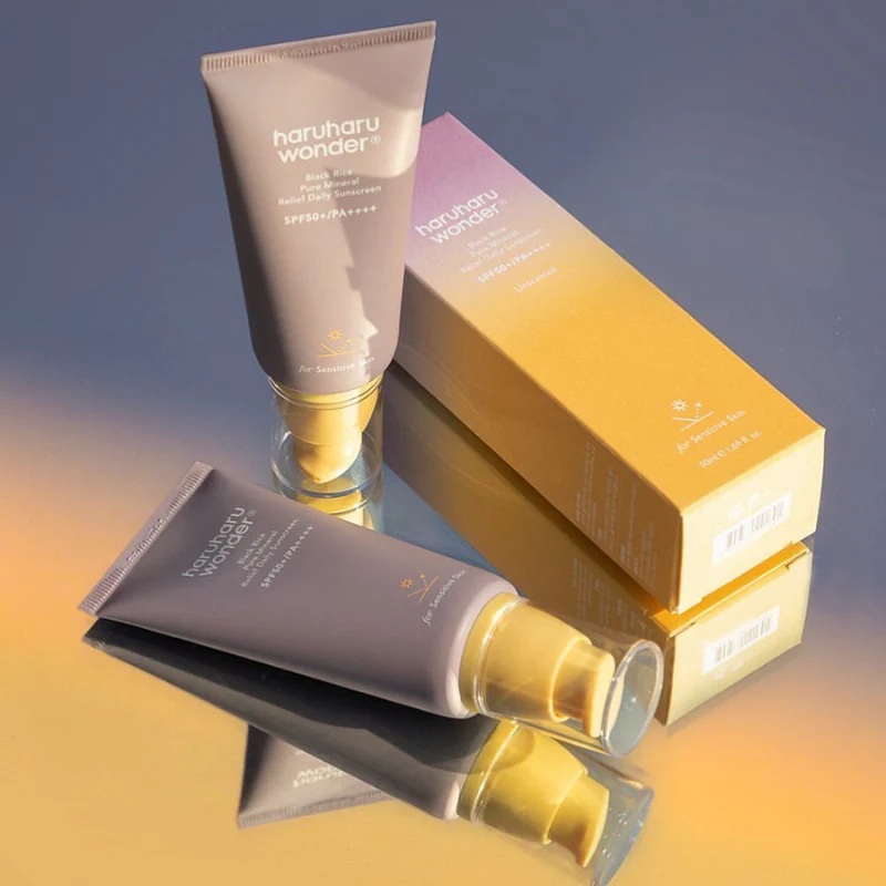 To tuber Haruharu Wonder Black Rice Pure Mineral Relief Daily Sunscreen SPF50+ PA++++ vises, en stående og en liggende, sammen med en gul og lilla produktboks på en reflekterende overflade, der ligner en stilfuld header-skabelon.