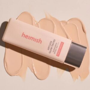 En tube Heimish - Artless Glow Tinted Sunscreen Shine Beige SPF50+ PA+++ står på en beige baggrund med cremede farveprøver af solcremen rundt om.