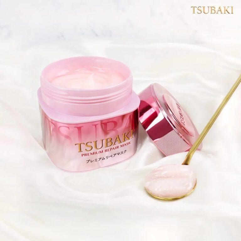 En åben krukke Shiseido - Tsubaki Premium Repair Mask Pink Camellia 180g står på en hvid marmoroverflade med låget af og en gylden ske med den lyserøde Camellia-infunderede formel ved siden af. Baggrunden er blød og elegant.