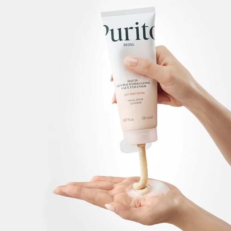 En hånd klemmer en tube med Purito SEOUL - Oat-In-Gentle Exfoliating Face Cleanser på en anden håndflade og uddeler beige, cremet rensemiddel med skum mod en hvid baggrund.