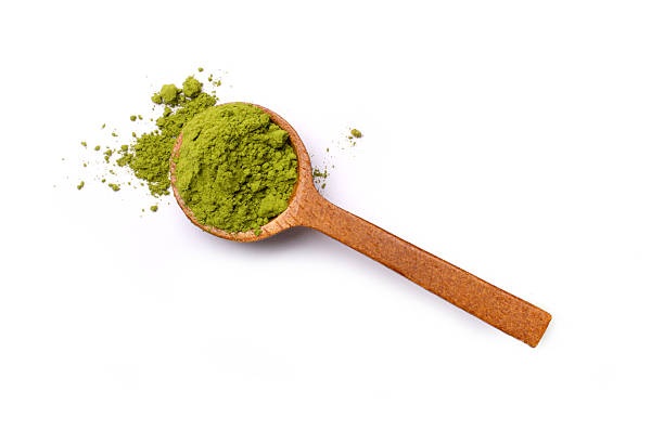 En træske fyldt med grønt pulver, sandsynligvis matcha eller et lignende tepulver, hviler ved siden af et lille spild på en hvid baggrund - perfekt til en henvisning til Ingrediensleksikon.