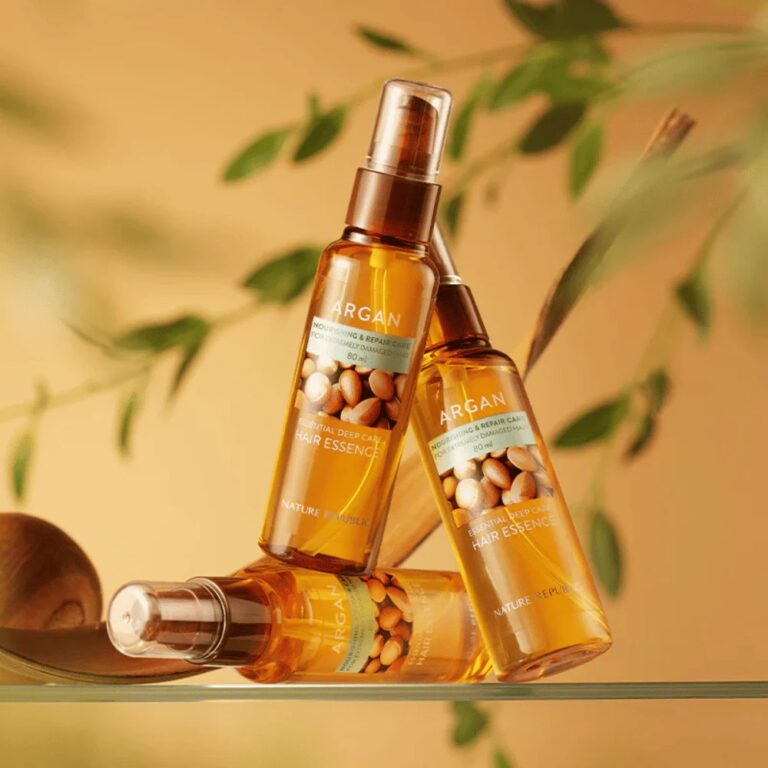 Tre flasker Nature Republic - Argan Essential Moisture Hair Oil er kunstnerisk arrangeret mod en gylden baggrund med slørede grønne blade, der understreger produktets nærende ingredienser til sundt hår.