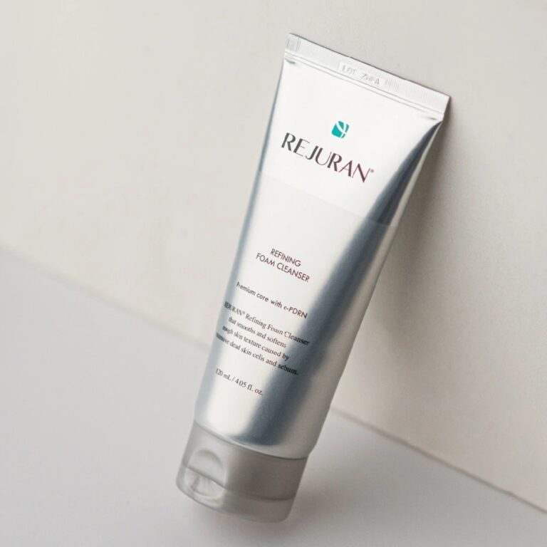 En sølv- og hvid tube Rejuran – Refining Foam Cleanser står oprejst på en lys overflade foran en hvid væg, hvor den gennemsigtige etiket og den gennemskinnelige hætte kan ses.