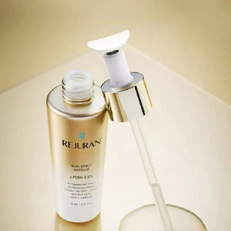 En guldfarvet flaske med etiketten »Rejuran – Dual Effect Ampoule« står åben på en skinnende beige overflade, mens hætten og den tilhørende dråbetæller ligger ved siden af.