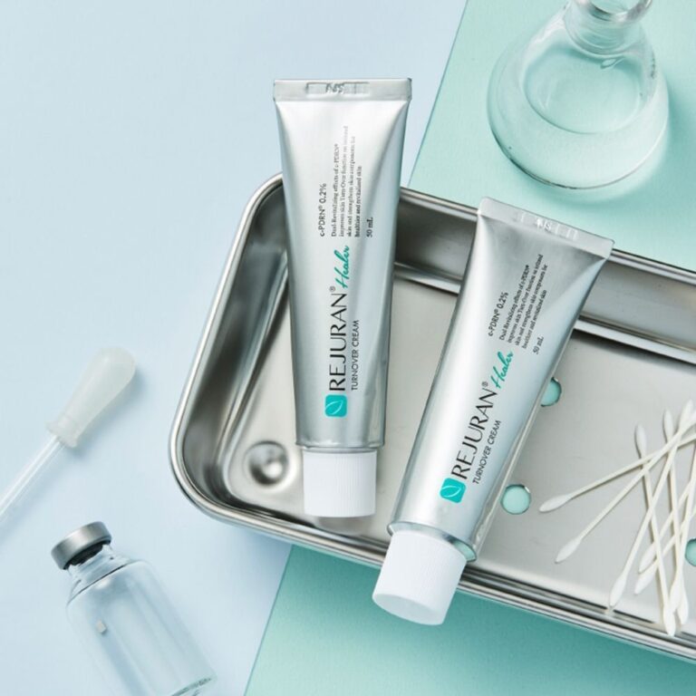 To sølvfarvede tuber med Rejuran – Turnover Cream Enhanced står på et metalfad sammen med vatpinde, en pipette og et lille glasflakon, anbragt på en lyseblå overflade for at give en forfinet hudplejeoplevelse.