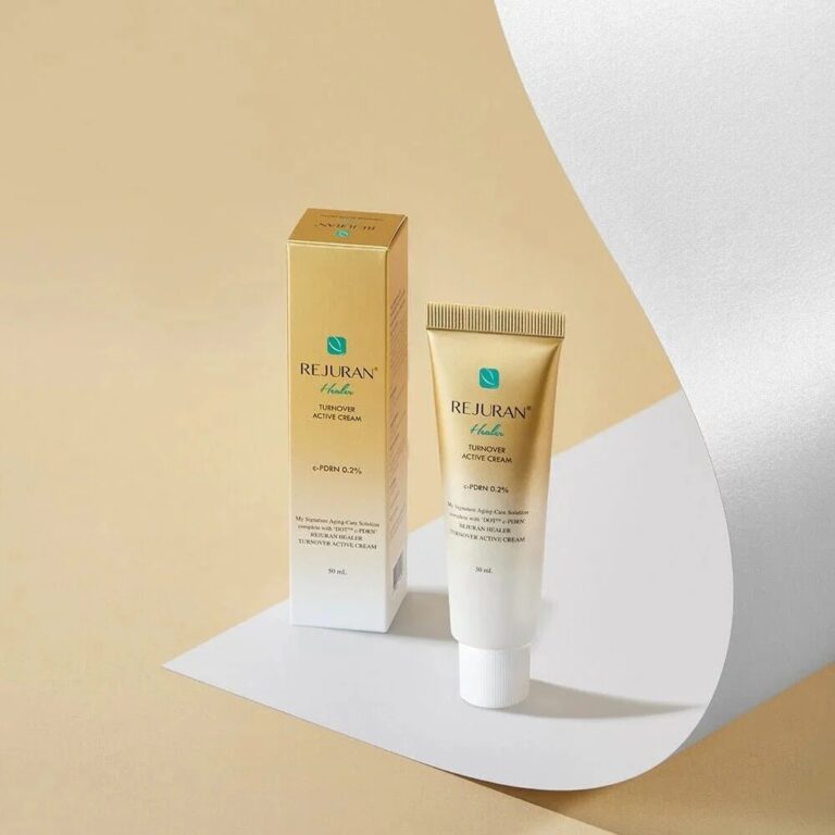 En guld- og hvid tube Rejuran – Active Cream Enhanced står ved siden af den tilhørende æske på en beige og hvid baggrund med et buet hvidt papirindslag.